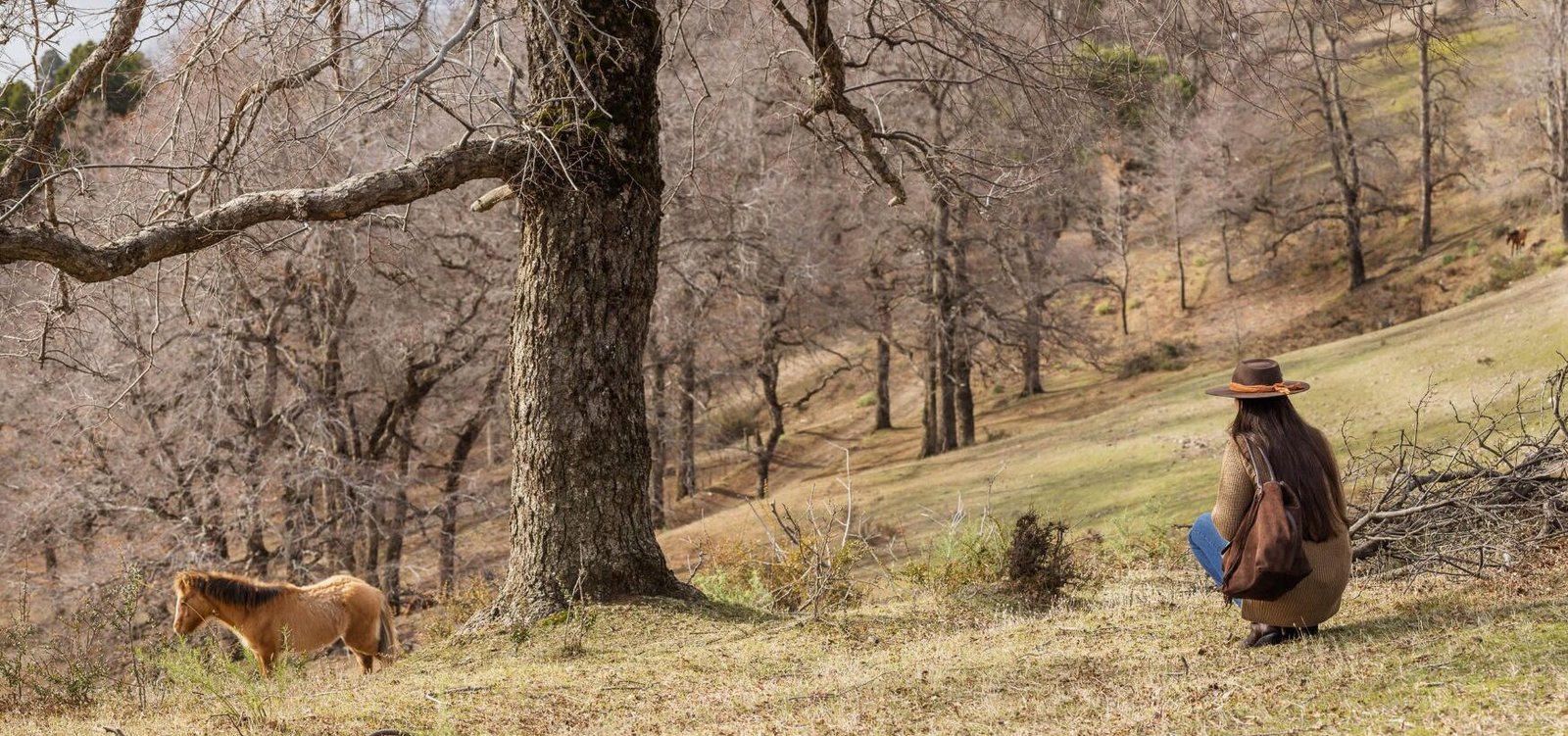 Mujer con sombrero y cartera de cuero con flecos junto a un caballo, en un bosque patagónico