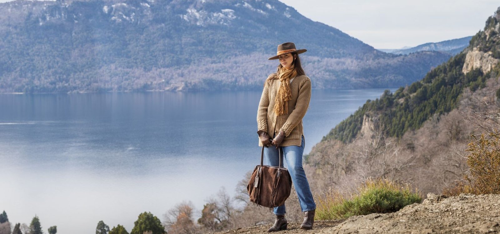 Mujer con cartera de cuero y botas marrones frente a un lago patagónico rodeado de montañas