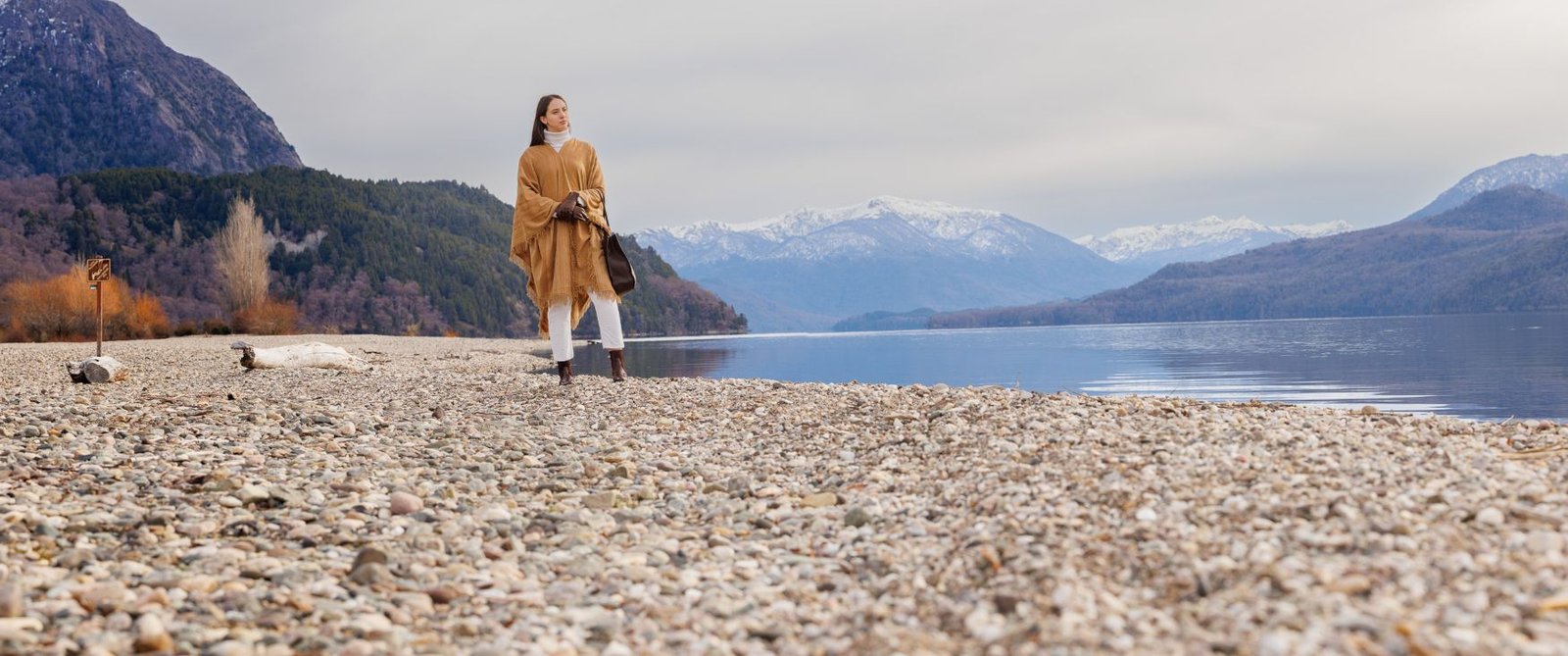 Mujer con poncho y cartera de cuero caminando por una playa de piedras patagónica, con montañas nevadas al fondo